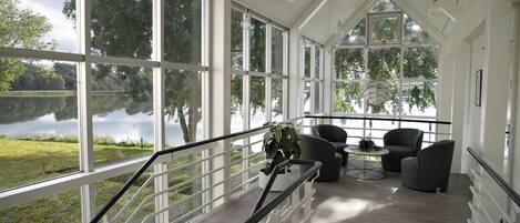 A spacious room with large windows overlooking a lake, a staircase with a glass railing, and modern furniture.