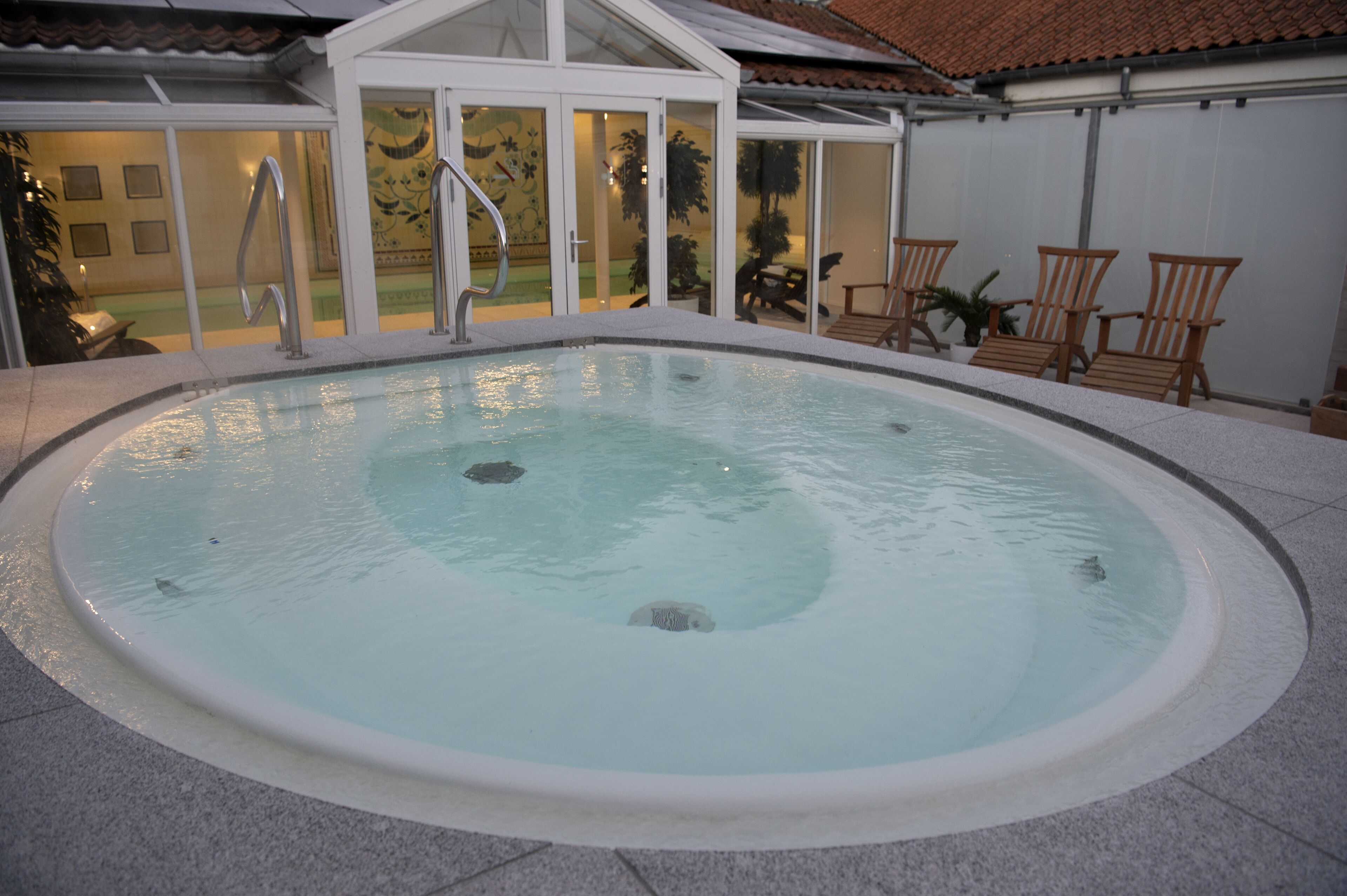 Ein runder Whirlpool mit gefliestem Rand, umgeben von einer Terrasse mit Holzstühlen und einer Glastür zu einem Innenbereich.