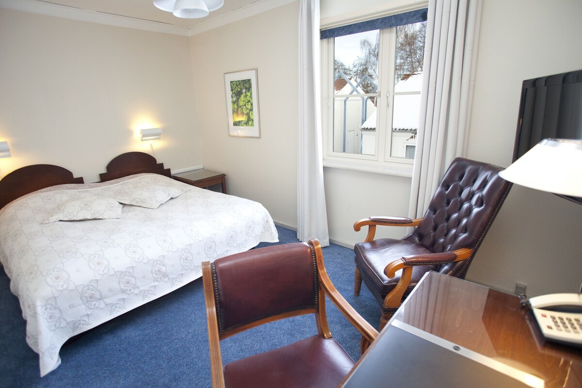 A hotel room with a bed, two bedside tables, a desk with a telephone, a leather chair, and a window with curtains.