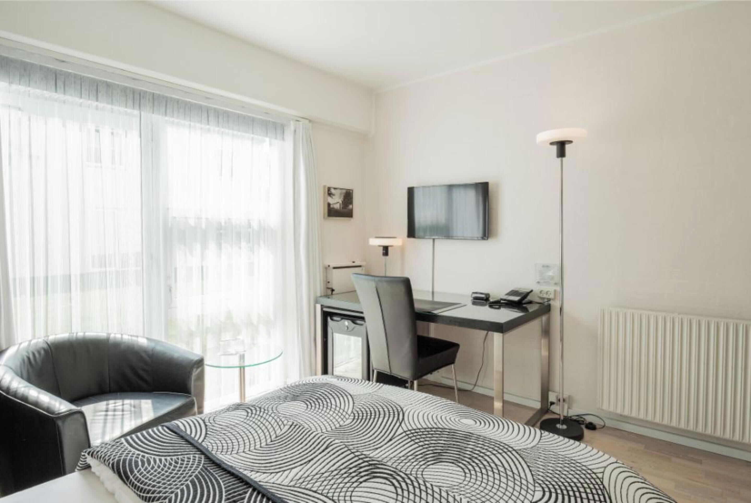 A modern hotel room with a bed, a desk, a chair, a TV, and a floor lamp.