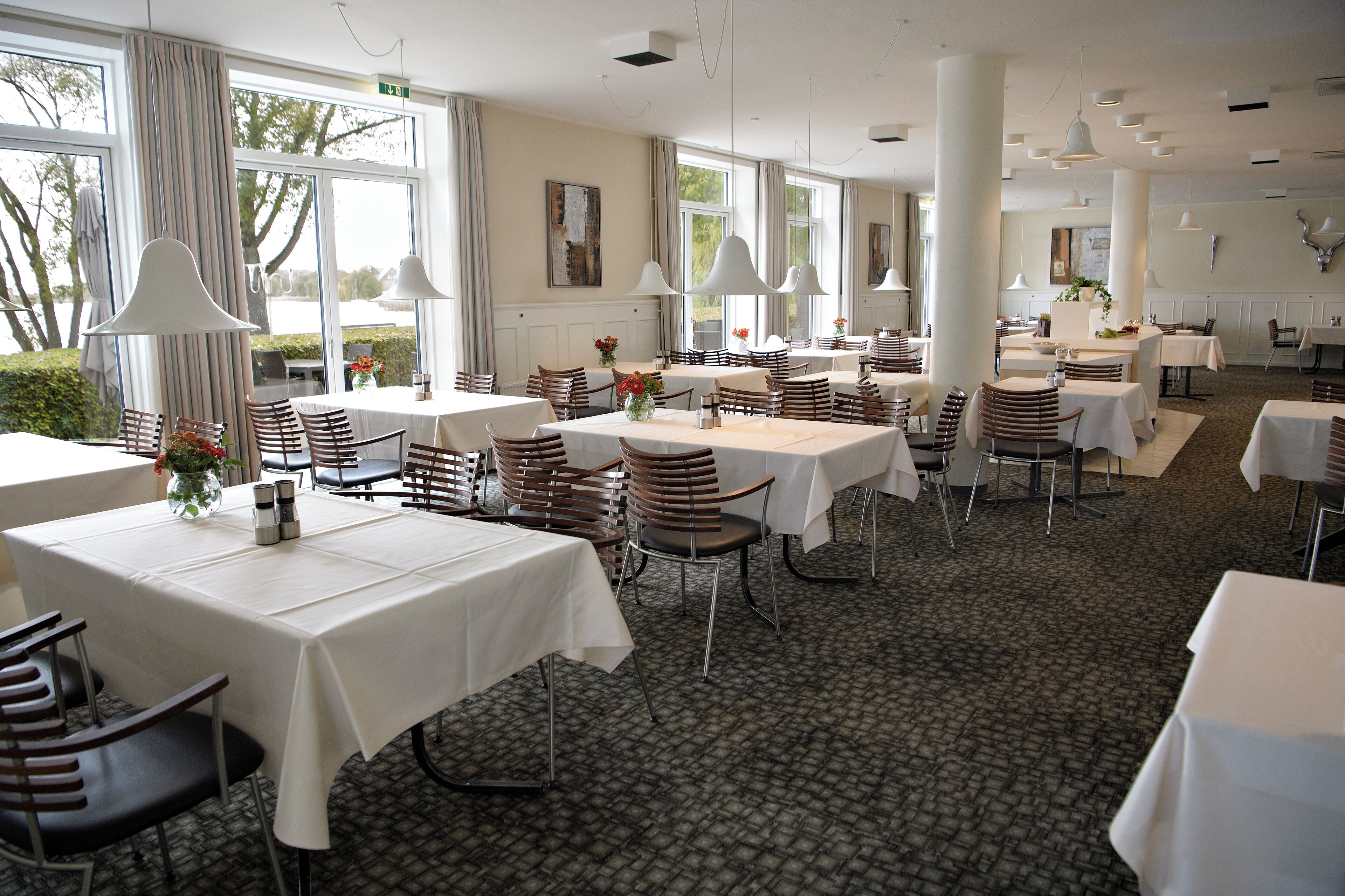 A spacious dining area with multiple tables and chairs, large windows, and pendant lighting.