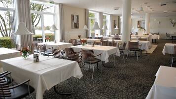 A spacious dining area with multiple tables and chairs, large windows, and pendant lighting.