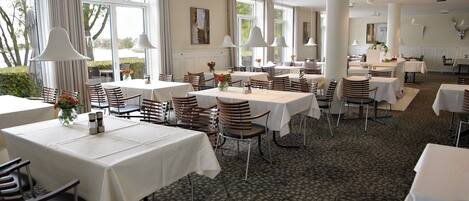 A spacious dining area with multiple tables and chairs, large windows, and pendant lighting.