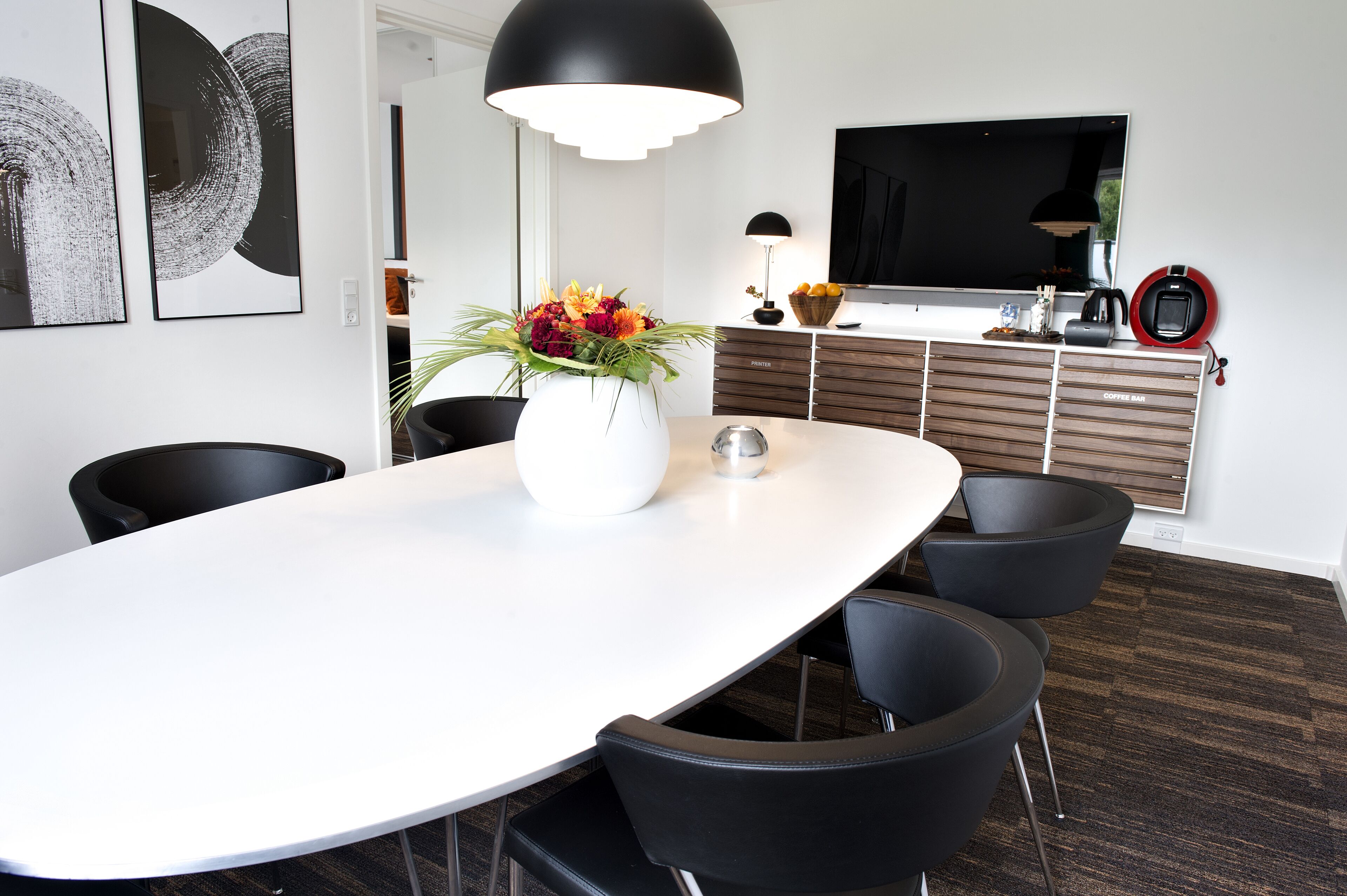 A modern dining area with a white table, black chairs, a vase of flowers, a large black pendant light, a flat-screen TV, and a coffee machine.