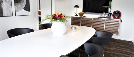 Un comedor moderno con una mesa blanca, sillas negras, un jarrón de flores, una lámpara colgante negra grande, un televisor de pantalla plana y una cafetera.