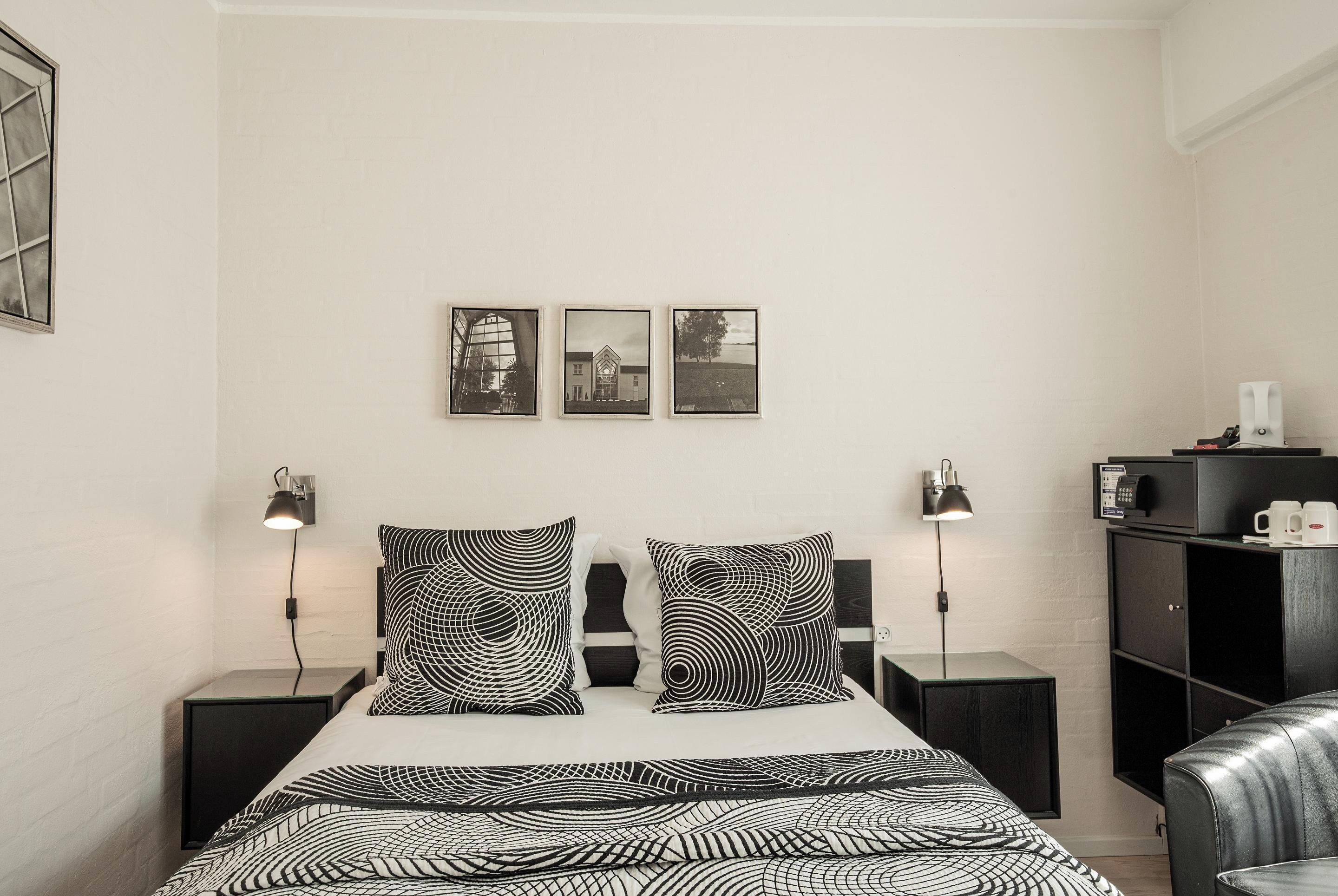 Une chambre moderne avec un lit, une literie à motifs noir et blanc, deux lampes de chevet, une armoire noire et trois tableaux encadrés accrochés au mur.