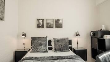 Une chambre moderne avec un lit, une literie à motifs noir et blanc, deux lampes de chevet, une armoire noire et trois tableaux encadrés accrochés au mur.
