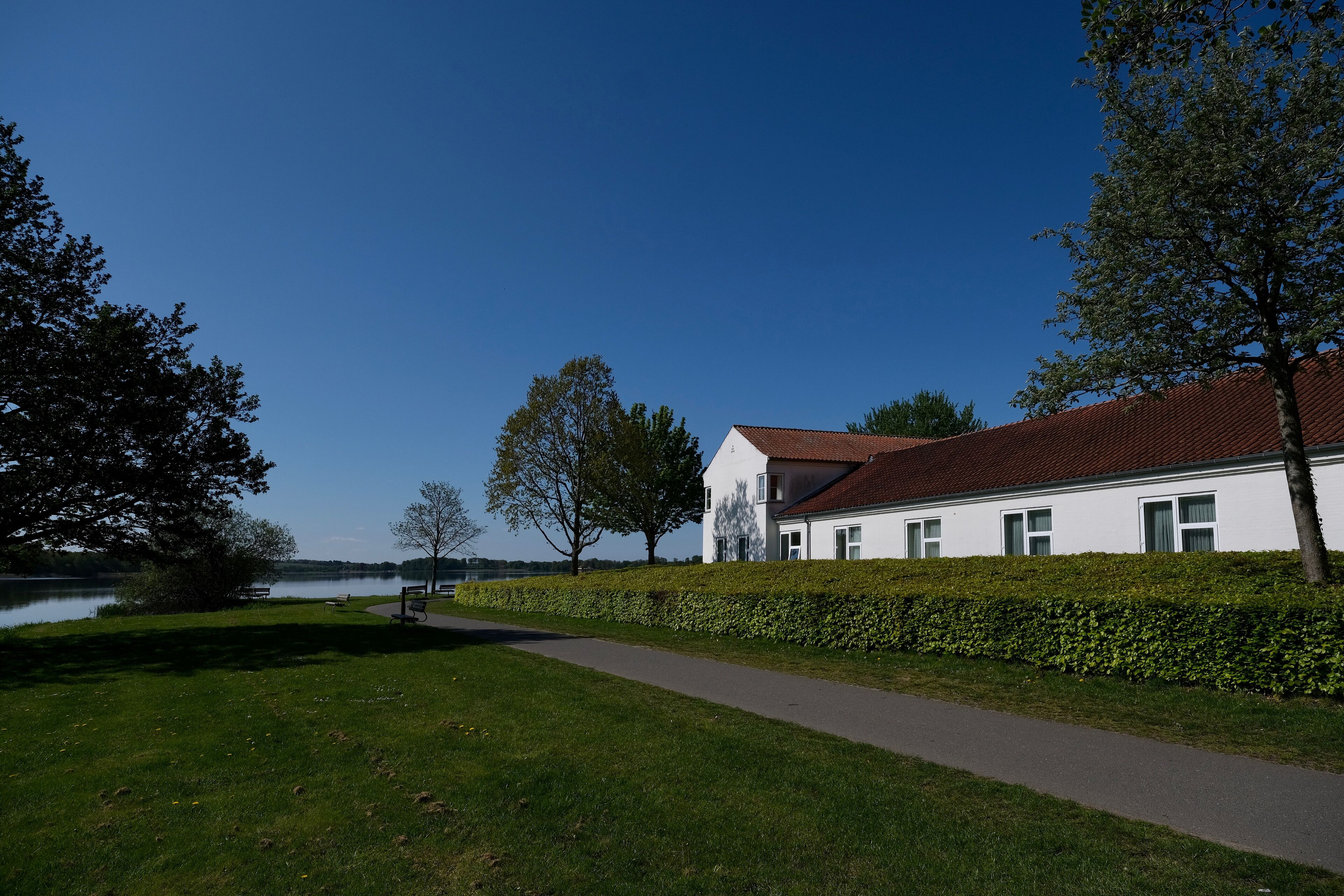 En hvid bygning med rødt tag, en anlagt sti og en sø i baggrunden.