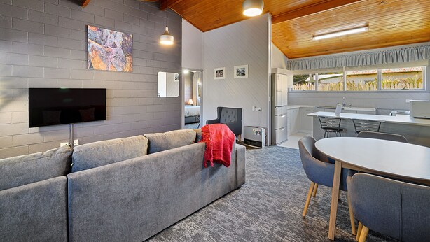 A modern living room with a grey sofa, a flat-screen TV, and a dining area with a round table and chairs. - Gold Rush Inn (Queenstown)