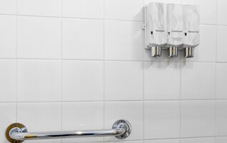 Baño con paredes de azulejos blancos, una barandilla de cromo y tres dispensadores etiquetados como 'Champú', 'Acondicionador' y 'Gel de ducha'.