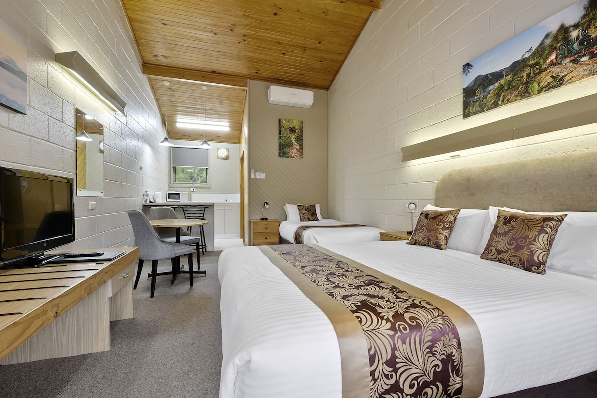 A hotel room with a wooden ceiling, a desk with a computer, a chair, two beds with decorative pillows, and a painting on the wall.