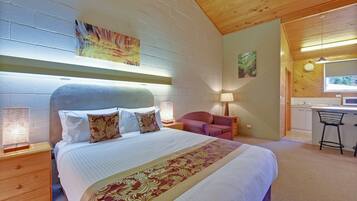 A bedroom with a large bed, a wooden headboard, a nightstand with a lamp, a chair, and a small kitchenette area.