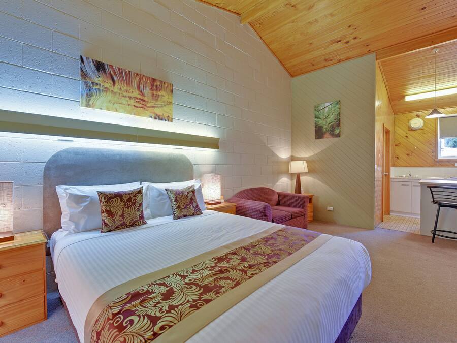 A bedroom with a large bed, a wooden headboard, a nightstand with a lamp, a chair, and a small kitchenette area.