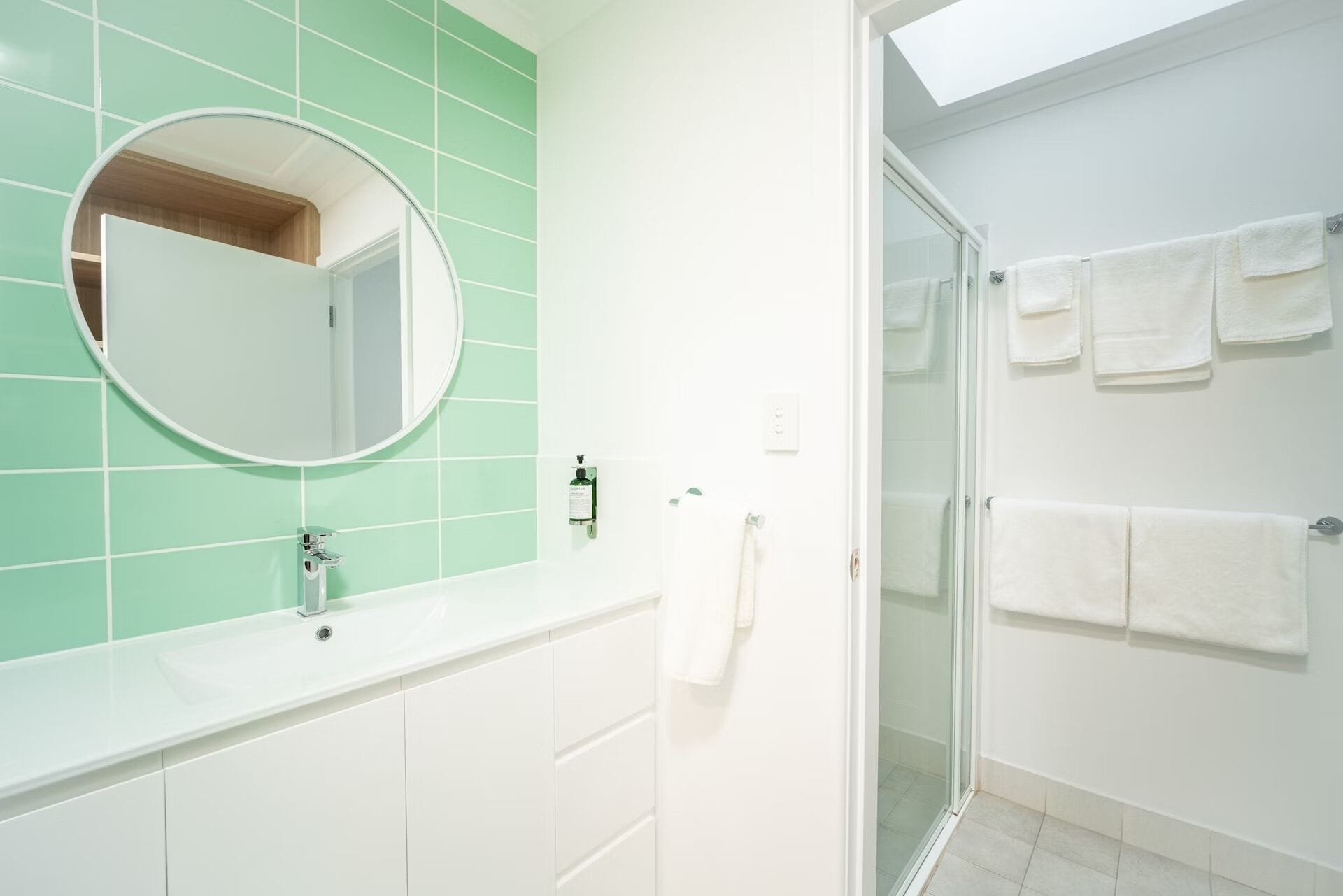 Une salle de bain avec un mur recouvert de carreaux verts, un miroir rond, un lavabo blanc et une cabine de douche en verre.