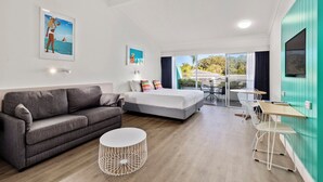 A modern hotel room with a grey sofa, a bed with colorful pillows, a white round side table, and a view of the outdoors through a sliding glass door. - The Cubana Resort Nambucca Heads (Nambucca Heads)