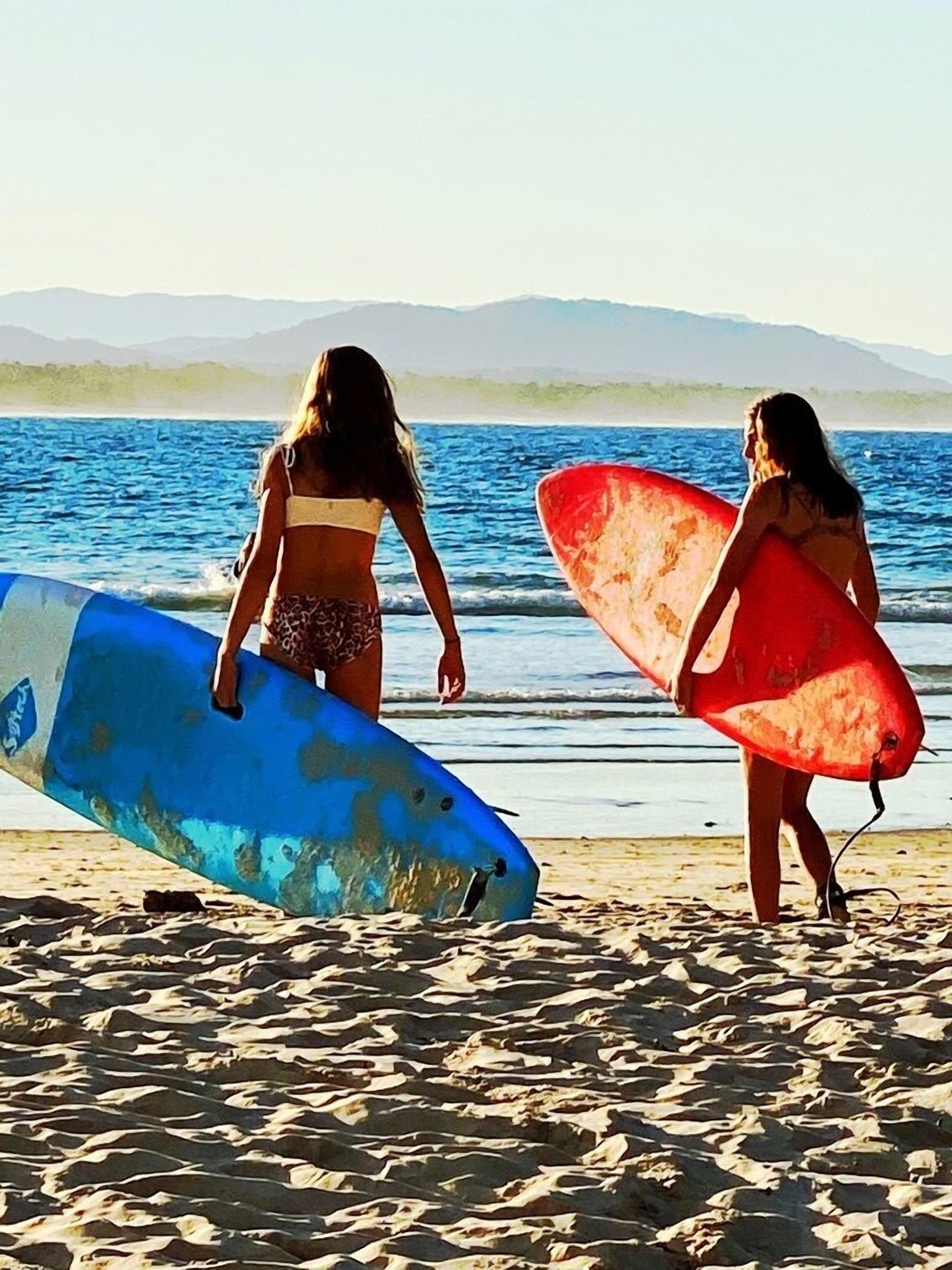 Two people carrying surfboards on a beach with a view of the sea and mountains in the background.