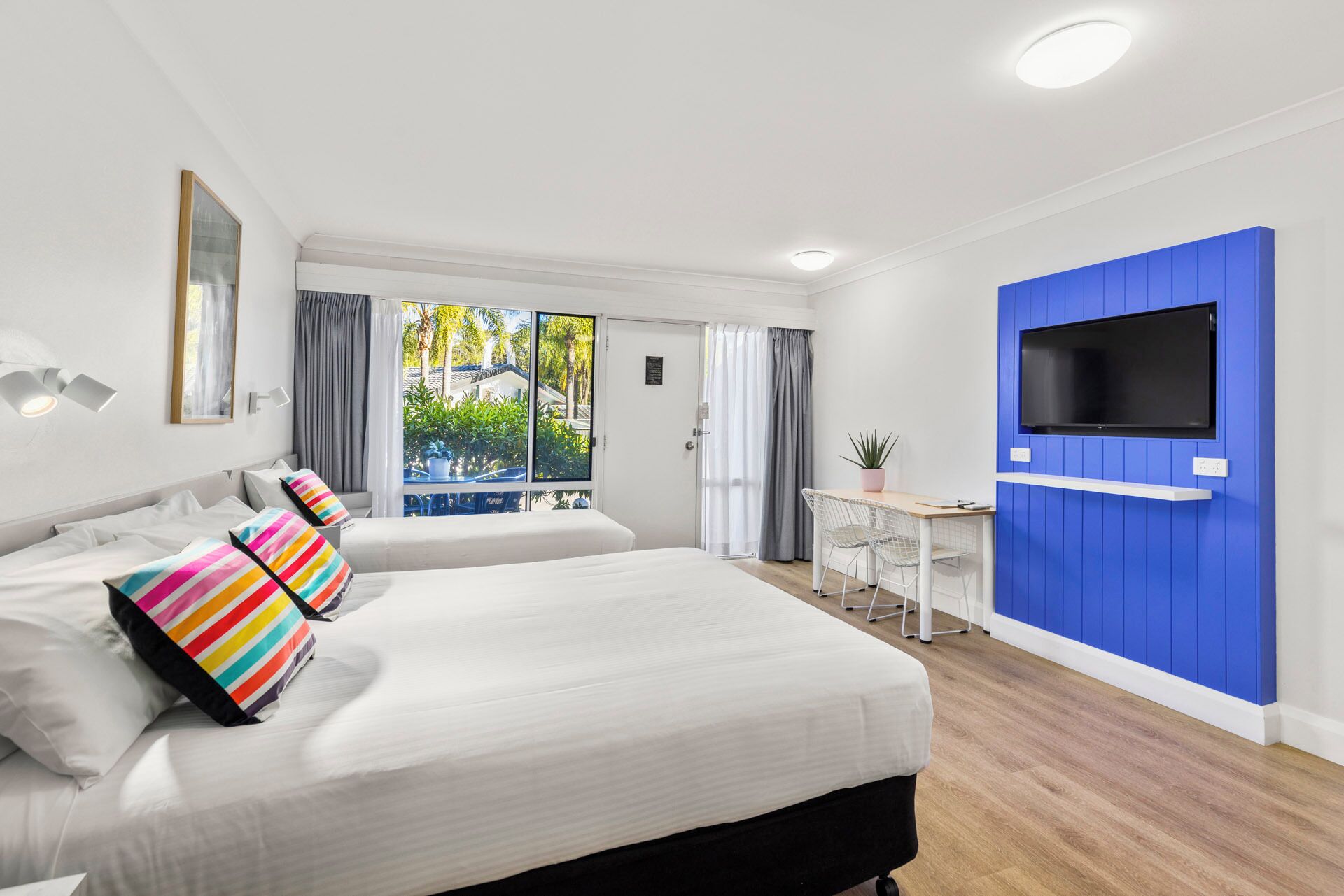 Una habitación de hotel moderna con dos camas, un escritorio, televisión y balcón con vistas a zonas verdes.