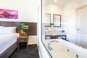 A hotel room with a bed, a nightstand with a vase of flowers, a large window, and a modern bathroom with a bathtub and two sinks.