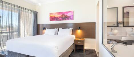 A hotel room with a large bed, a nightstand with a lamp, a bathroom with a sink and mirror, and a painting on the wall.
