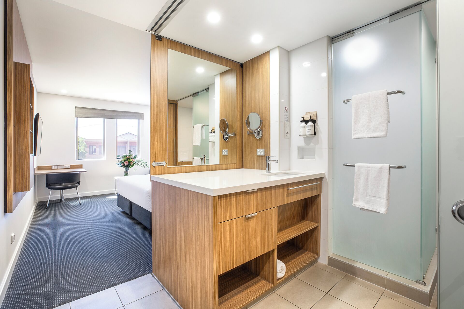 Un baño moderno con tocador de madera, un espejo grande, cabina de ducha de vidrio y una pequeña zona de estar con escritorio.