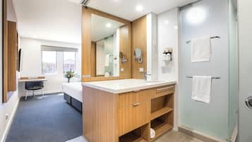 A modern bathroom with a wooden vanity, a large mirror, a glass shower enclosure, and a small seating area with a desk.