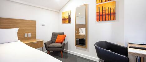 A modern hotel room with a large bed, a desk with a chair, a seating area with a chair and a sofa, and artwork on the walls.