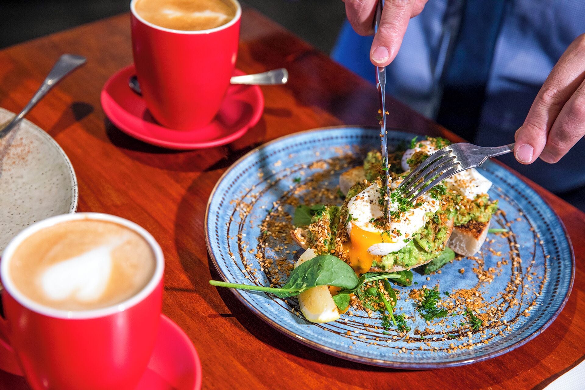 A person dining at a table with a blue plate containing a meal, a red cup of coffee, and a spoon.