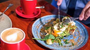 Eine Person isst an einem Tisch, auf dem sich ein blaues Teller mit einer Mahlzeit, eine rote Kaffeetasse und ein Löffel befinden.