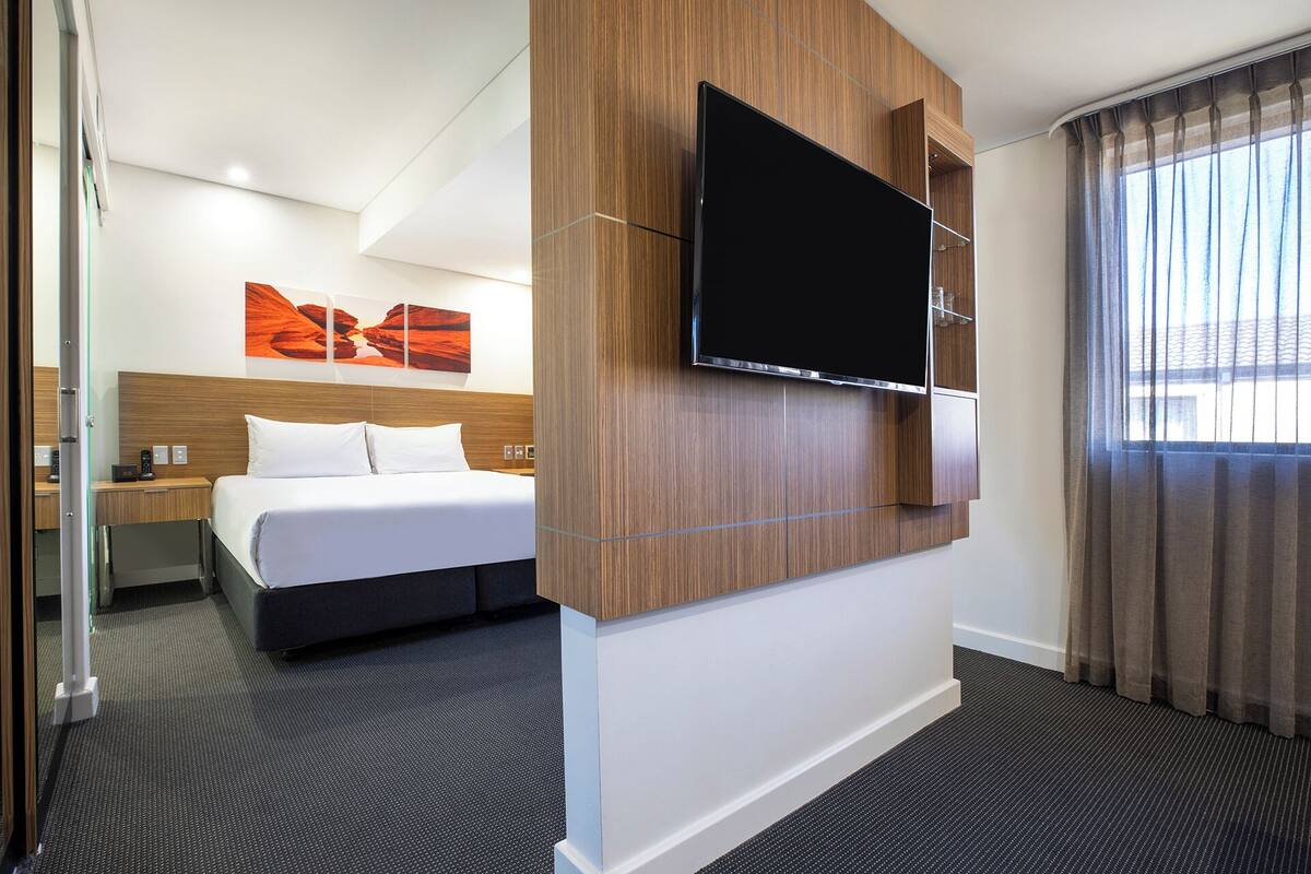 Habitación de hotel con cama, televisor de pantalla plana en la pared, mesita de noche y ventana con cortinas.