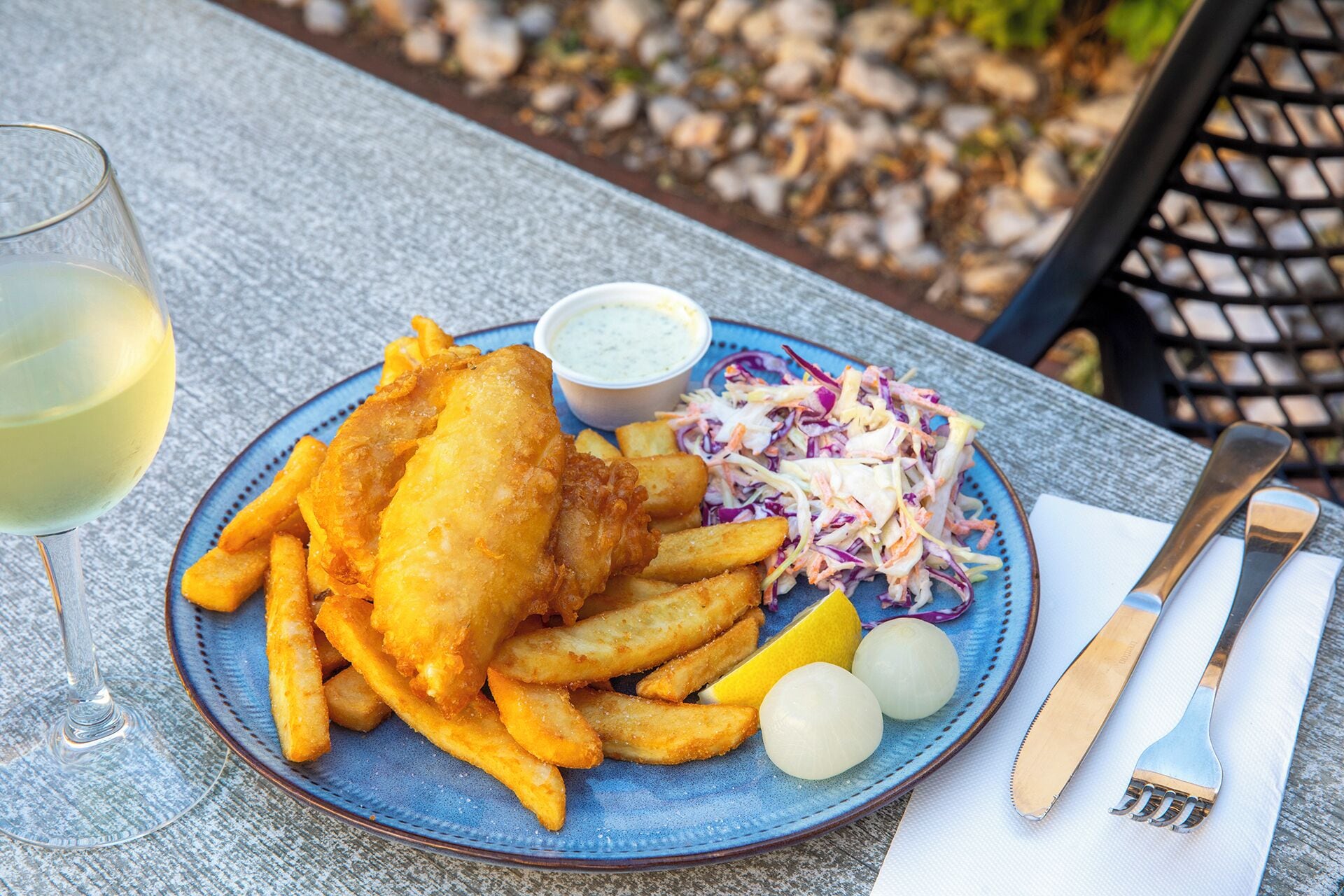 A plate of fish and chips with a side of coleslaw and a glass of white wine on a table.