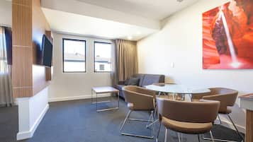 Una habitación de hotel moderna con un sofá, dos sillones, una mesita y un televisor fijado a un panel de madera.