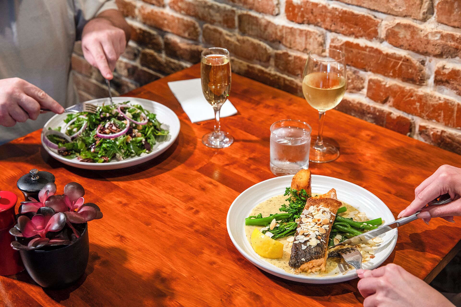 A wooden table with two plates of food, a glass of water, and a glass of wine.