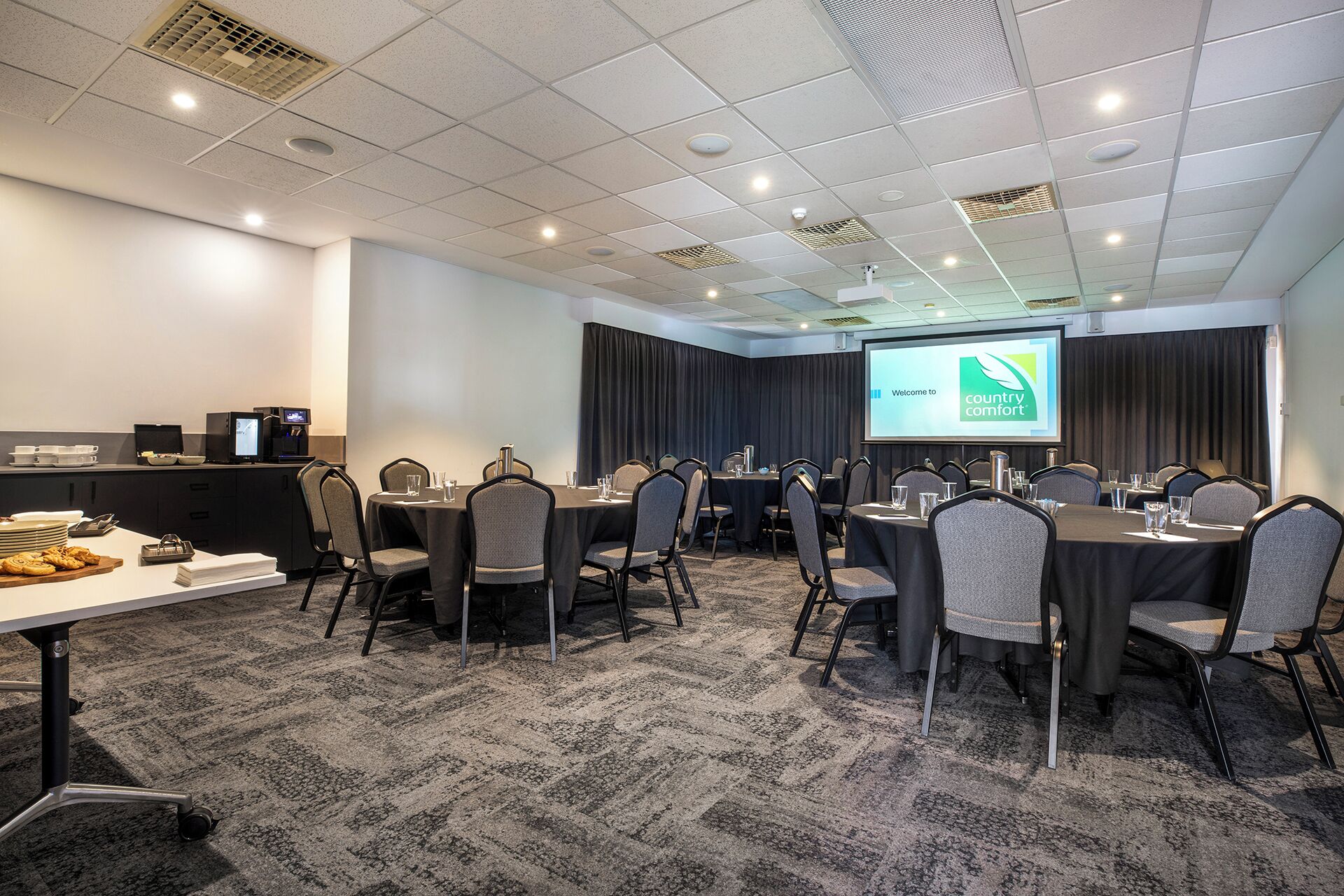 A conference room with multiple tables and chairs, a projector screen displaying a welcome message, and a buffet table with food items.