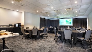 A conference room with multiple tables and chairs, a projector screen displaying a welcome message, and a buffet table with food items.
