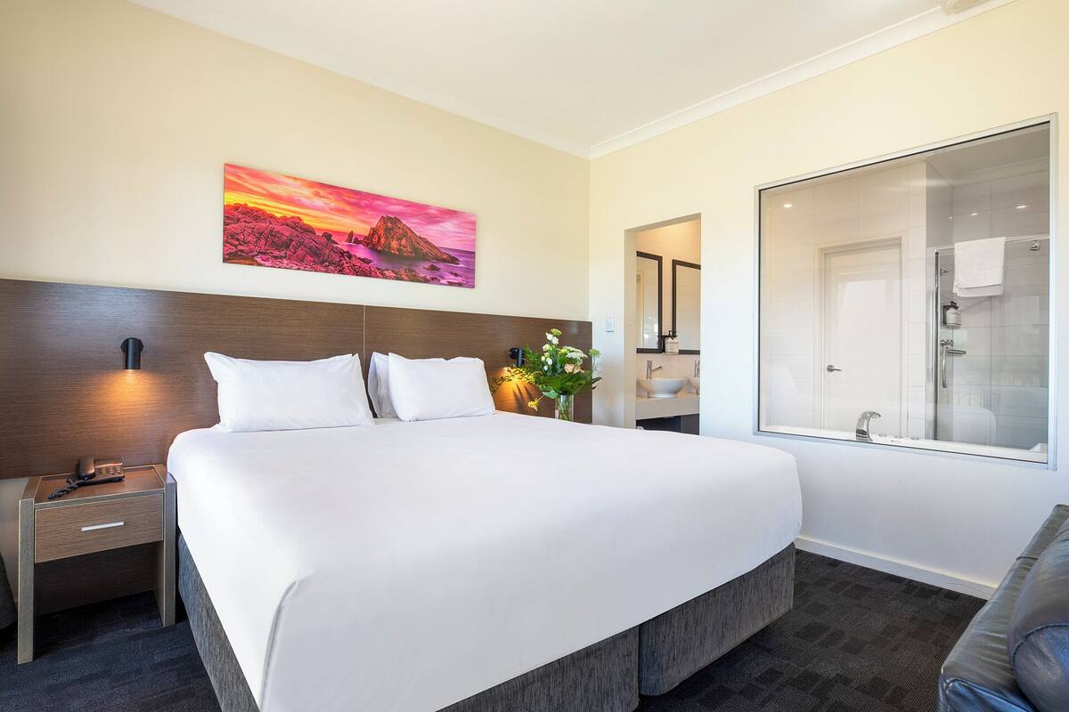 Habitación de hotel con una cama grande, una mesita de noche con teléfono, un cuadro de un atardecer, un baño con ducha de vidrio y un espejo.