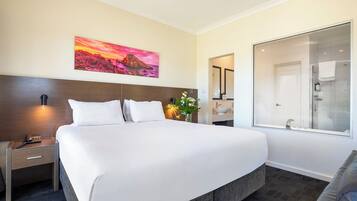 Habitación de hotel con una cama grande, una mesita de noche con teléfono, un cuadro de un atardecer, un baño con ducha de vidrio y un espejo.