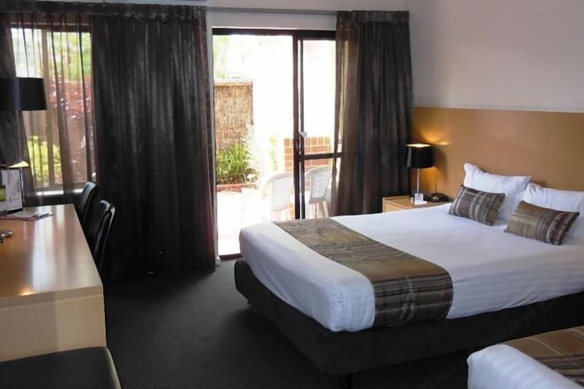 A hotel room with a large bed, a desk, and a chair. There is a window with curtains, a lamp, and a view of the outside.