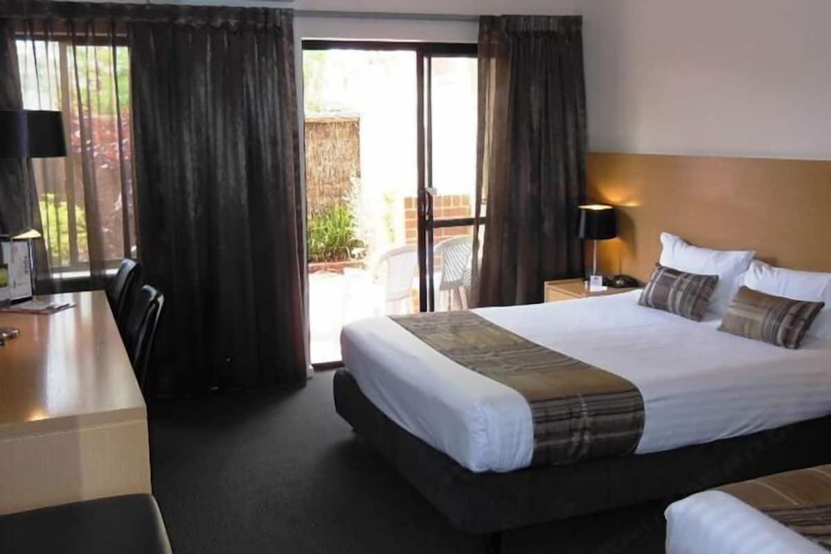A hotel room with a large bed, a desk, and a chair. There is a window with curtains, a lamp, and a view of the outside.