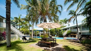 A poolside area with a table and chairs, surrounded by palm trees and a building with a balcony. - Mango House Resort (Jubilee Pocket)