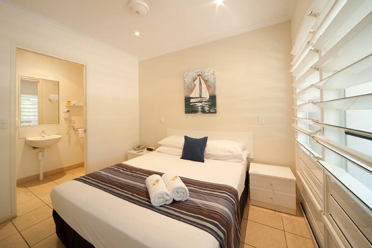 Une chambre d’hôtel avec un lit, une table de chevet, une commode, un miroir, un tableau représentant un voilier et une fenêtre avec des stores.