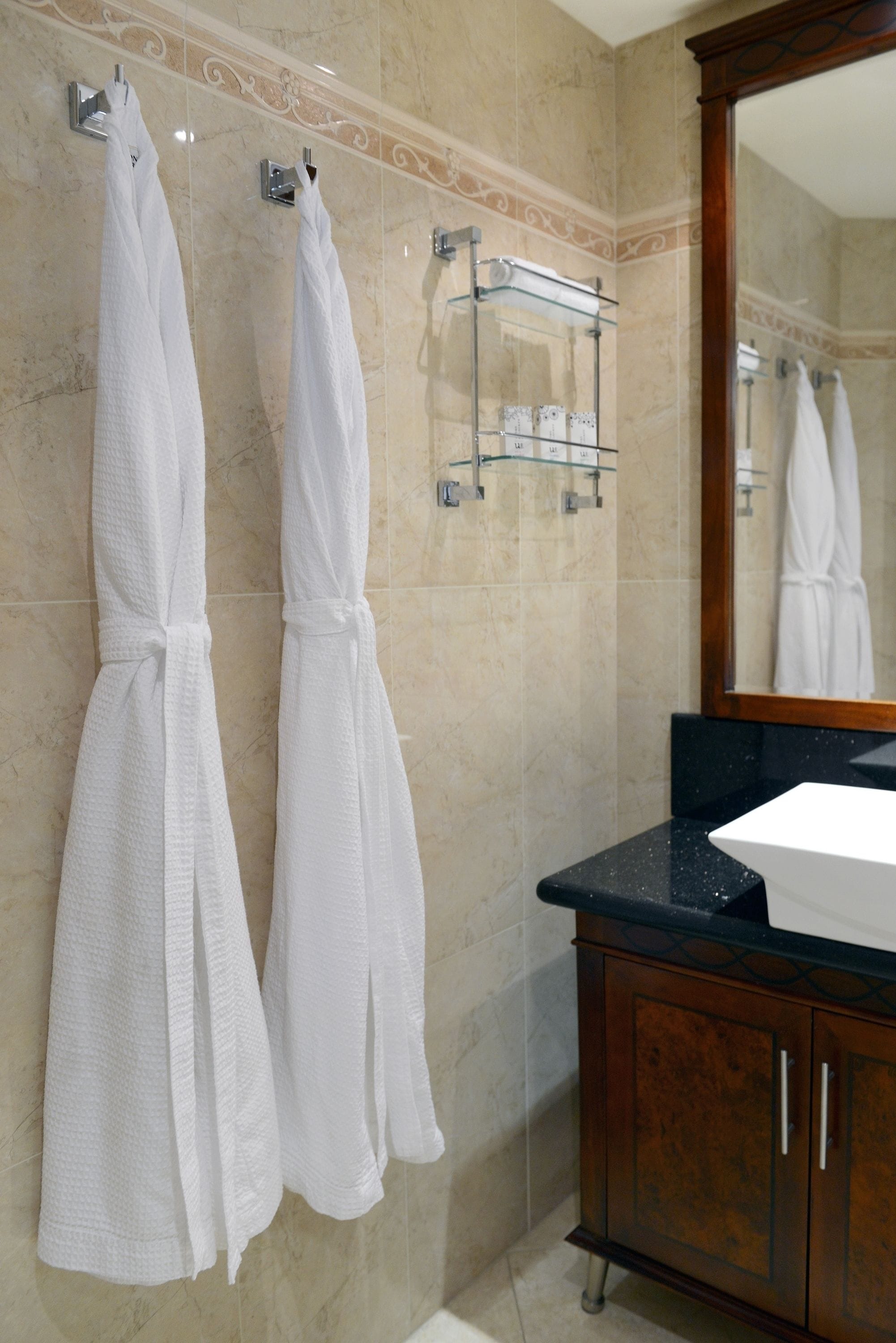 A bathroom with two white robes hanging on hooks, a glass shelf with toiletries, and a wooden cabinet with a sink.