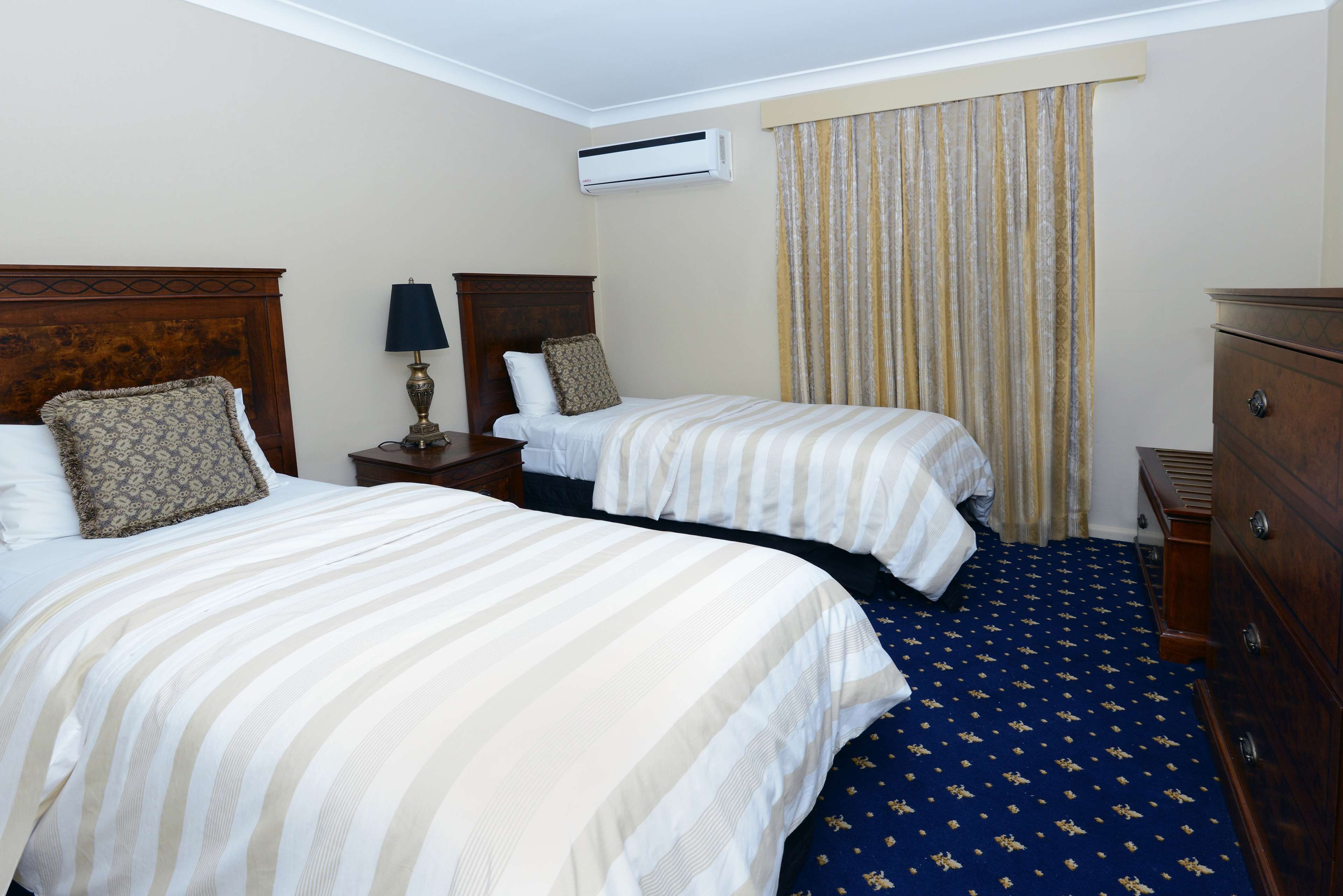 Quarto de hotel com duas camas, uma mesa de cabeceira com abajur, cômoda e janela com cortinas.