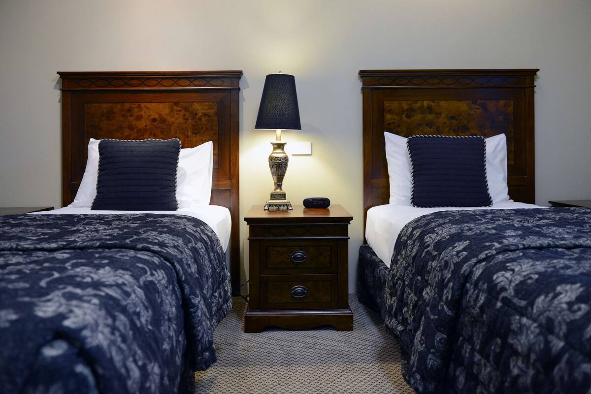 A hotel room with two beds, each with a dark blue patterned bedspread and white pillows. There is a wooden nightstand between the beds with a lamp on it.