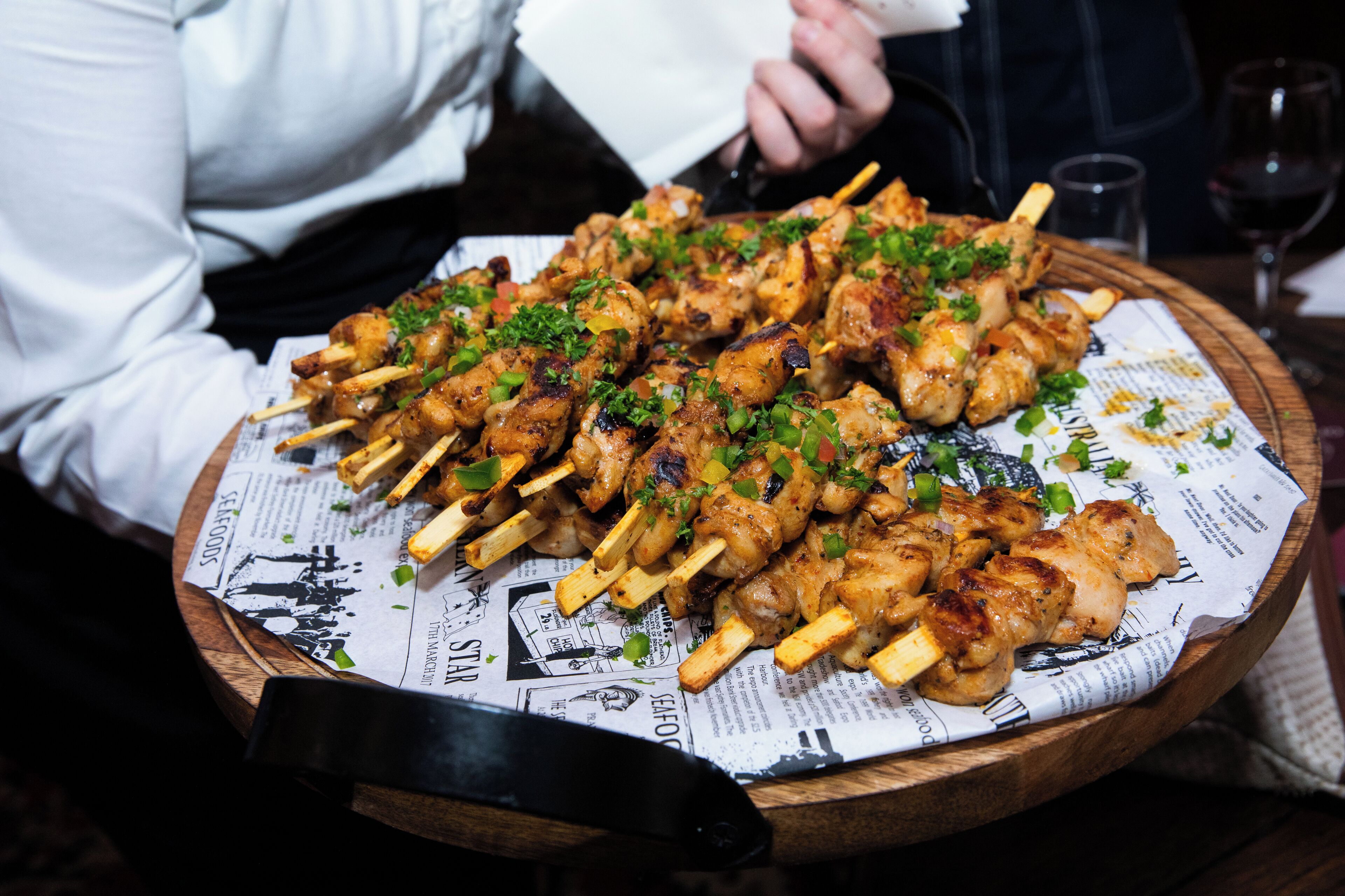 A wooden tray with skewers of grilled chicken garnished with herbs, placed on a newspaper-lined surface.