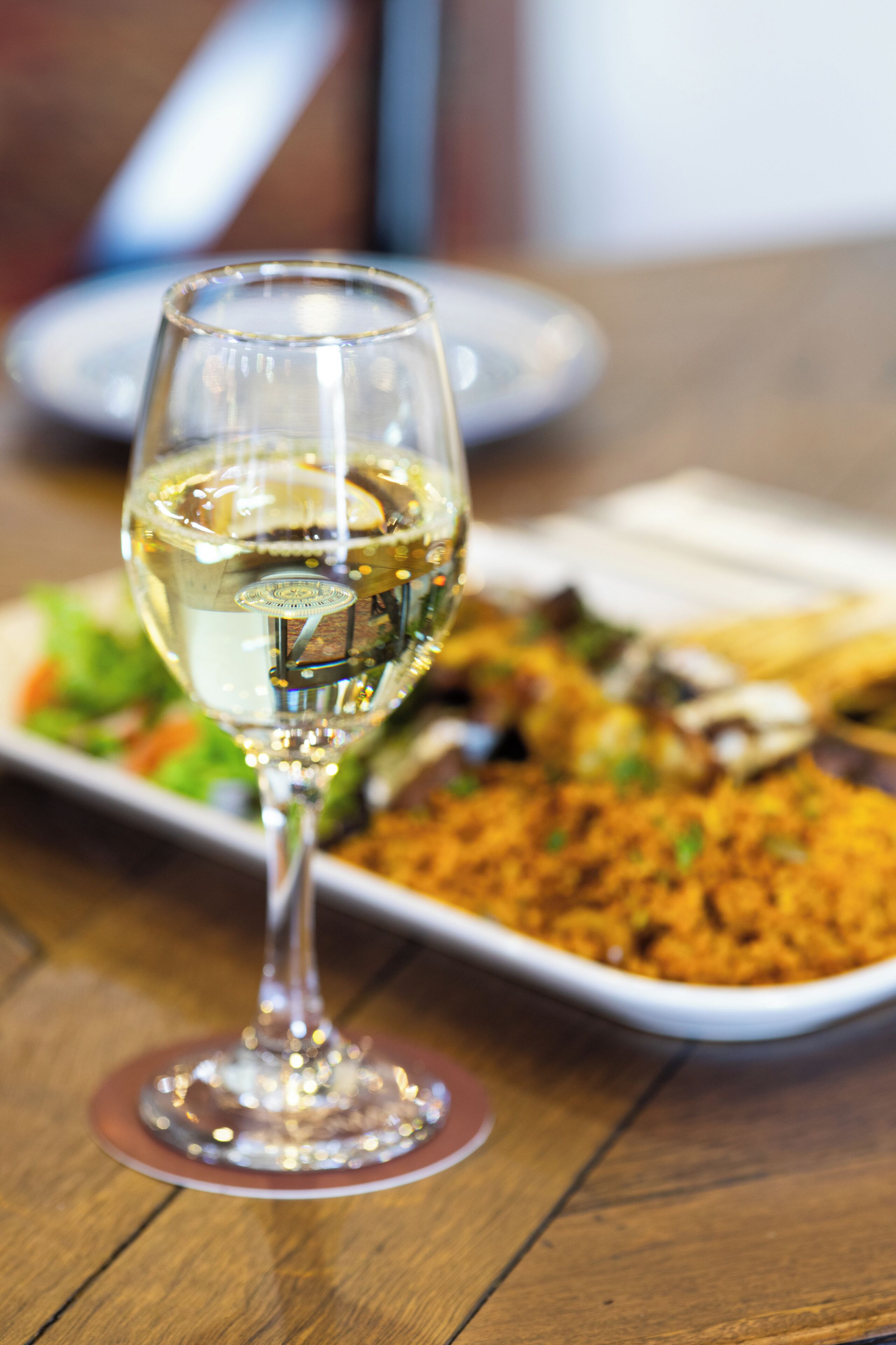 Una copa de vino blanco sobre una mesa de madera, con un plato de comida al fondo.