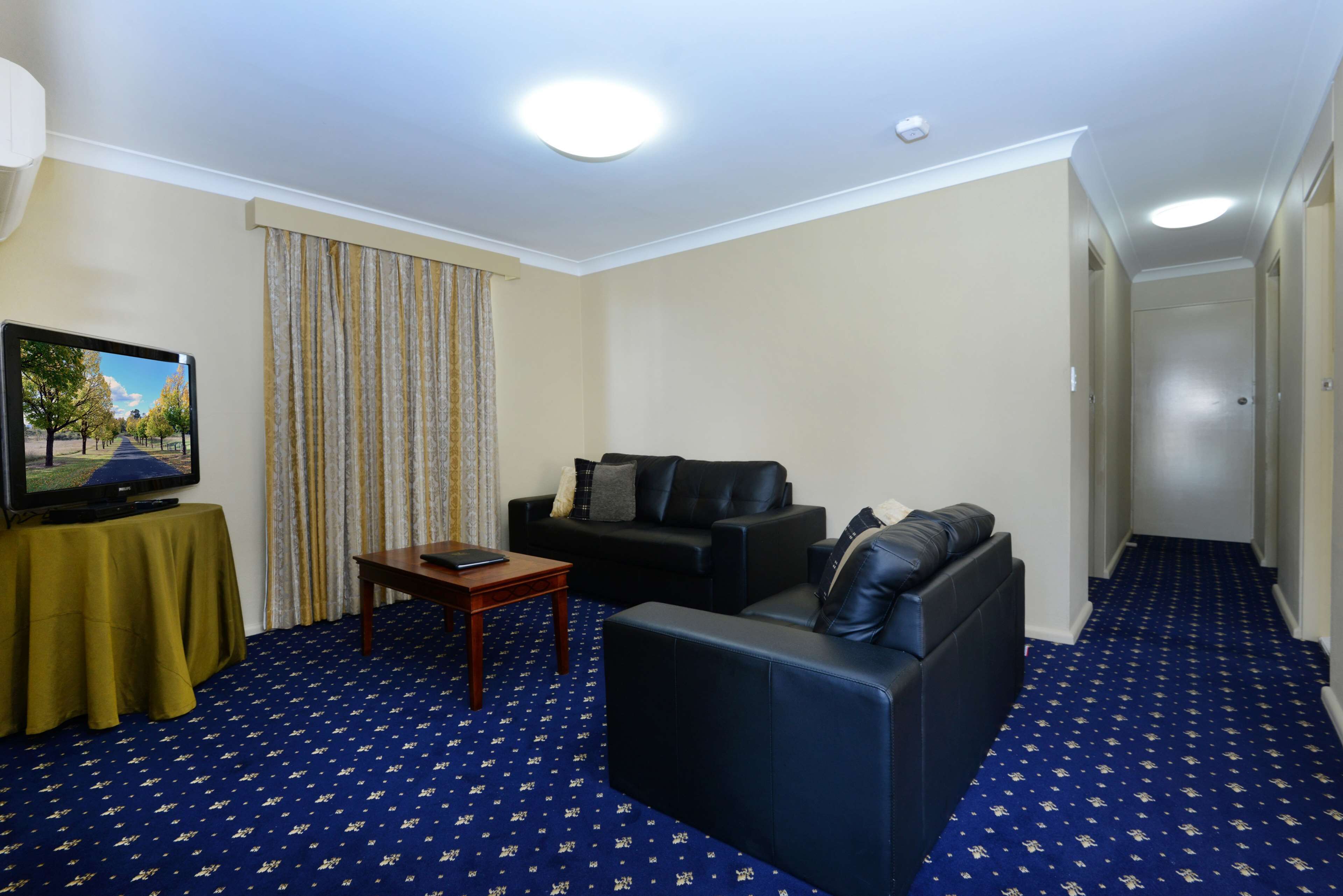 A hotel room with a blue carpet, two black sofas, a wooden coffee table, a television on a small table, and a curtain.