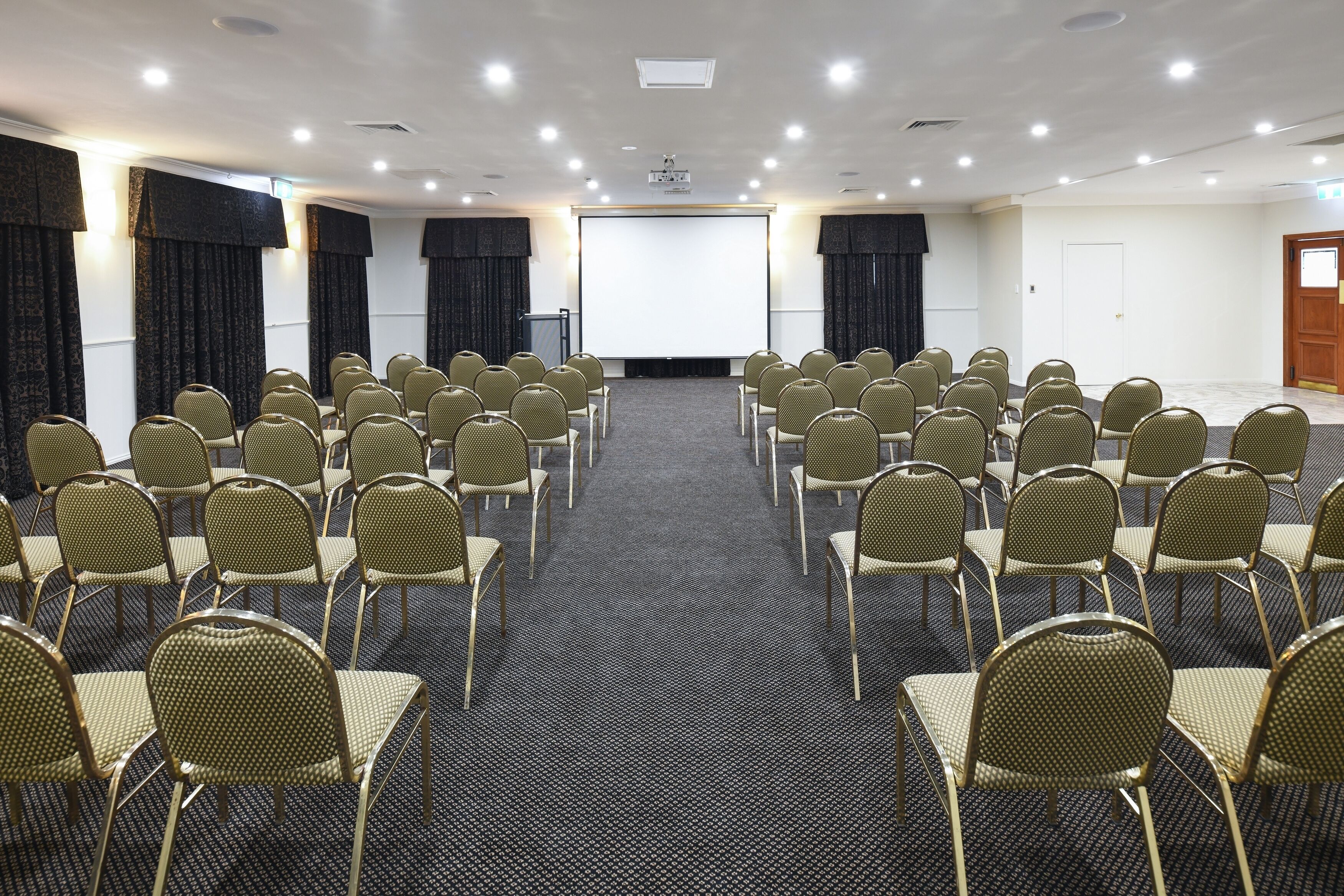 A conference room with rows of chairs facing a large screen, carpeted floor, and curtains on the walls.