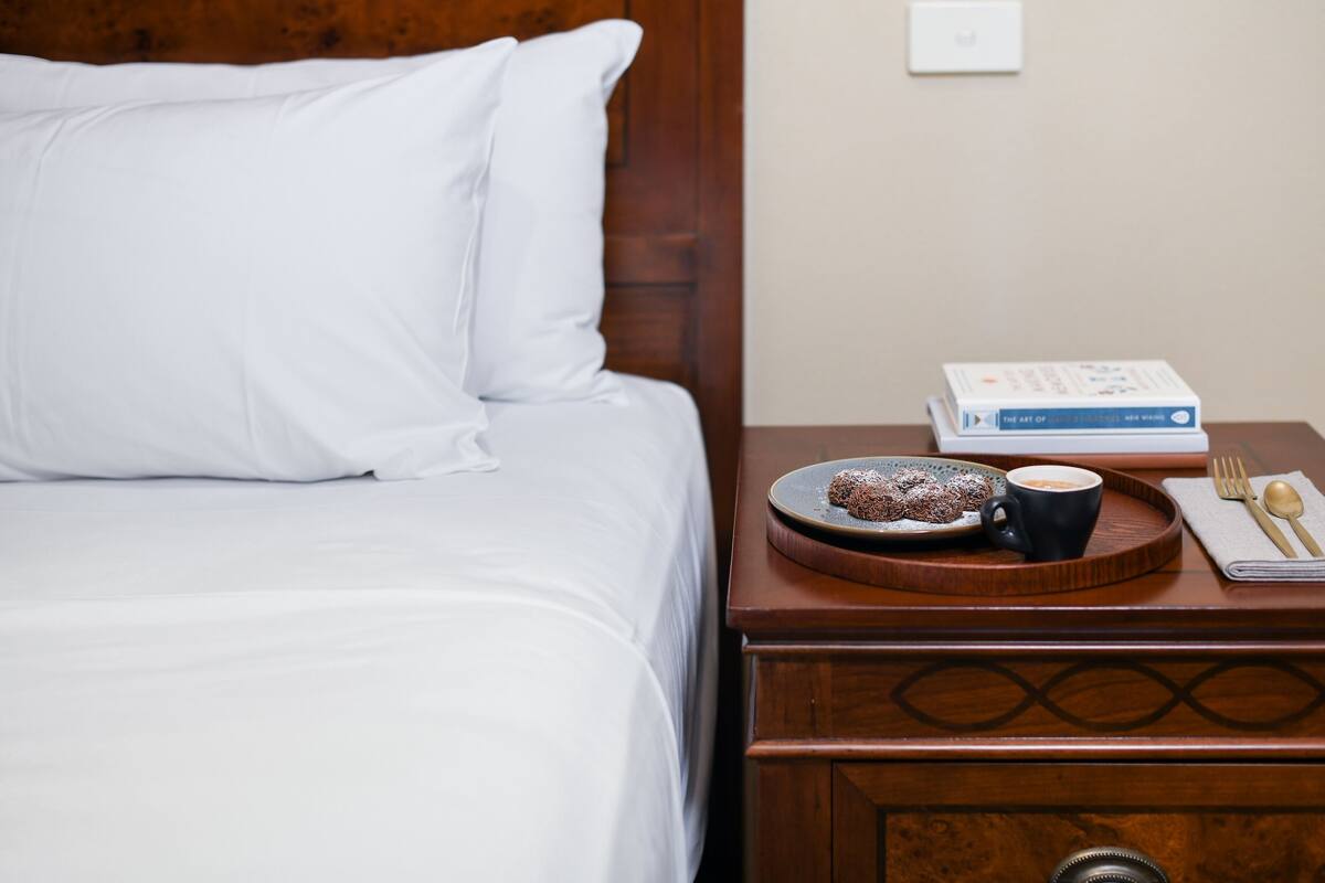 A neatly made bed with white linens, a bedside table with a book, a plate of food, a cup of coffee, and a set of cutlery.