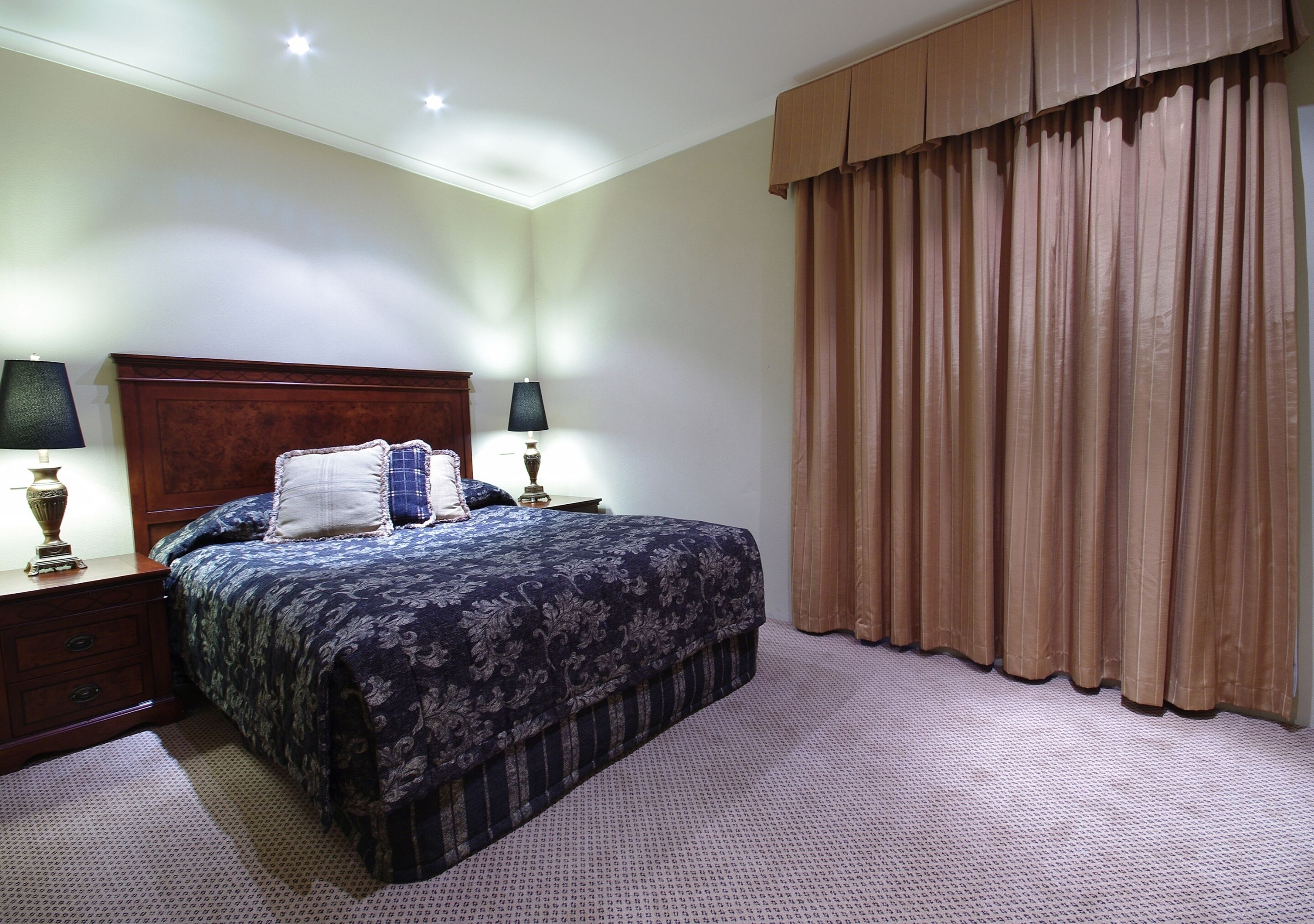 A neatly made bed with a dark patterned bedspread, two bedside lamps, a wooden nightstand, and a large window with brown curtains.