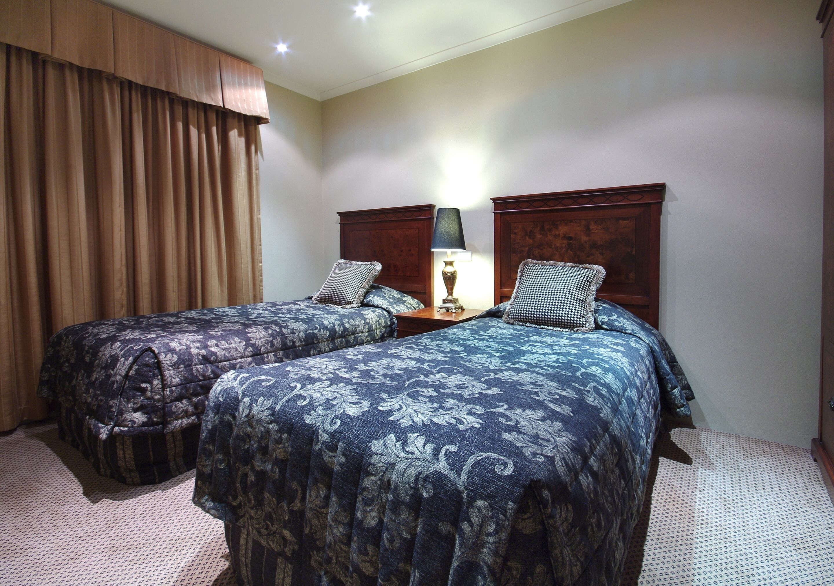 A hotel room with two beds, each with a patterned bedspread, a nightstand with a lamp, and a wooden headboard.